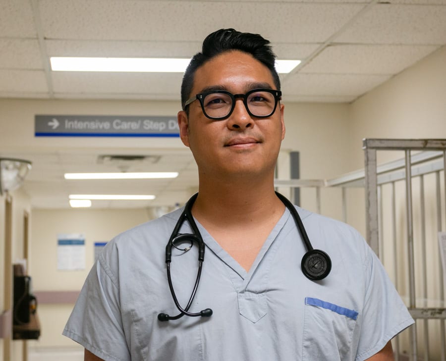 A young Asian male doctor wearing scrubs, a stethoscope and stylish black glasses smiles for the camera.