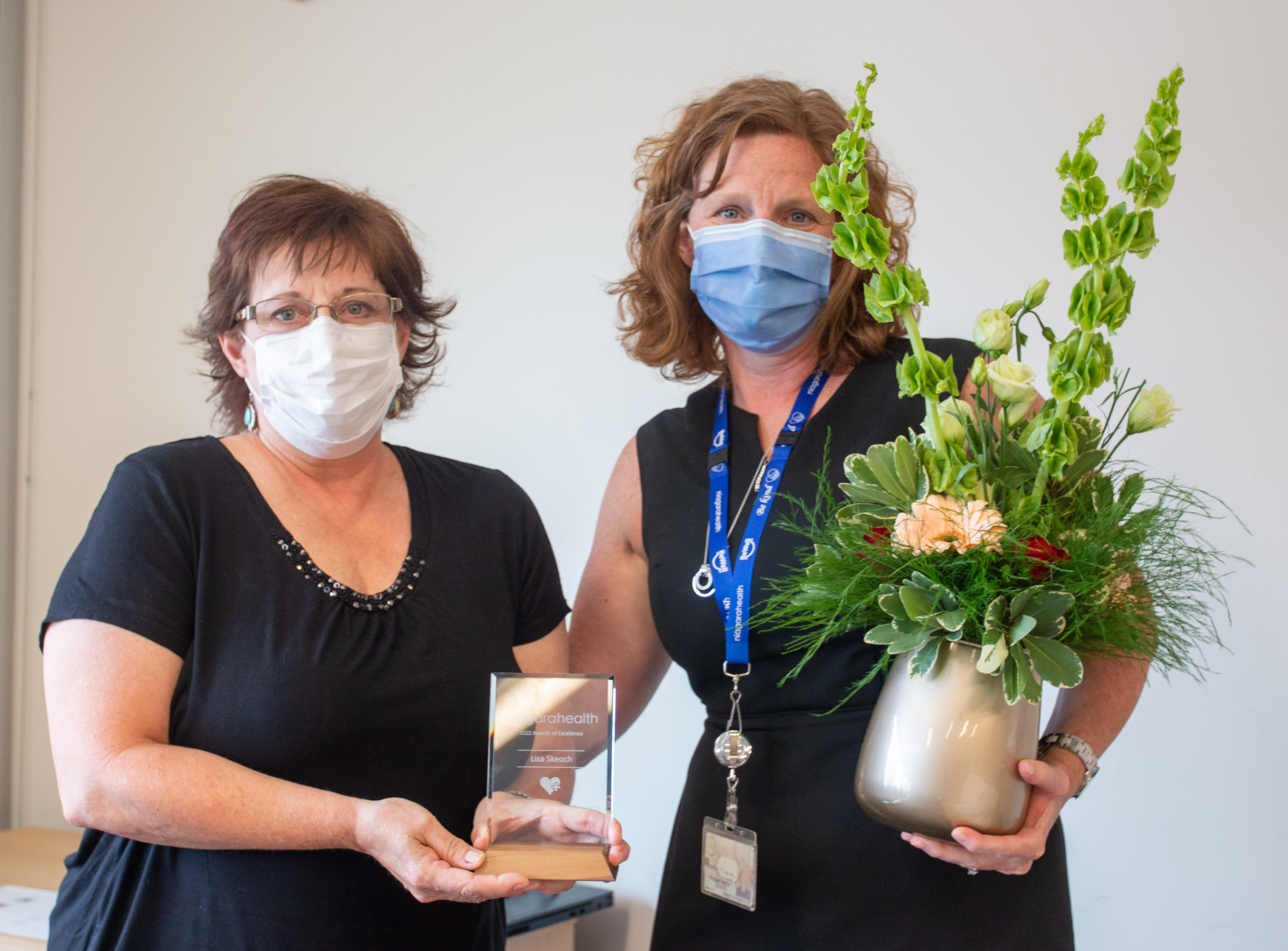 Two women stand together. One holds an award, the other holds flowers. They are wearing masks and smiling.