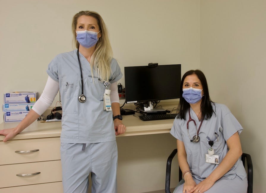 Two nurses dressed in scrubs lean against a desk with a computer on it.