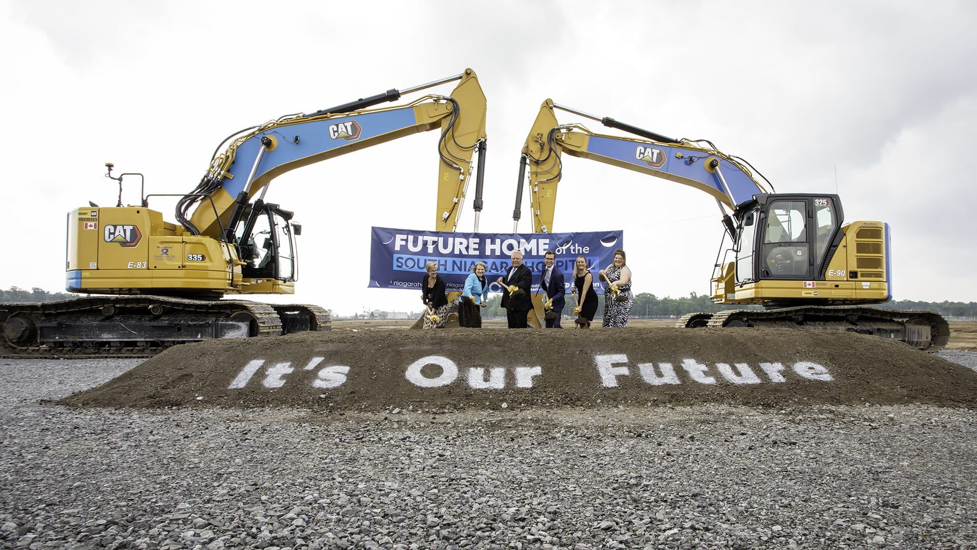 A group of people gather around a pile of dirt with ceremonial shovels and two backhoes behind them. A sign reads It's Our Future