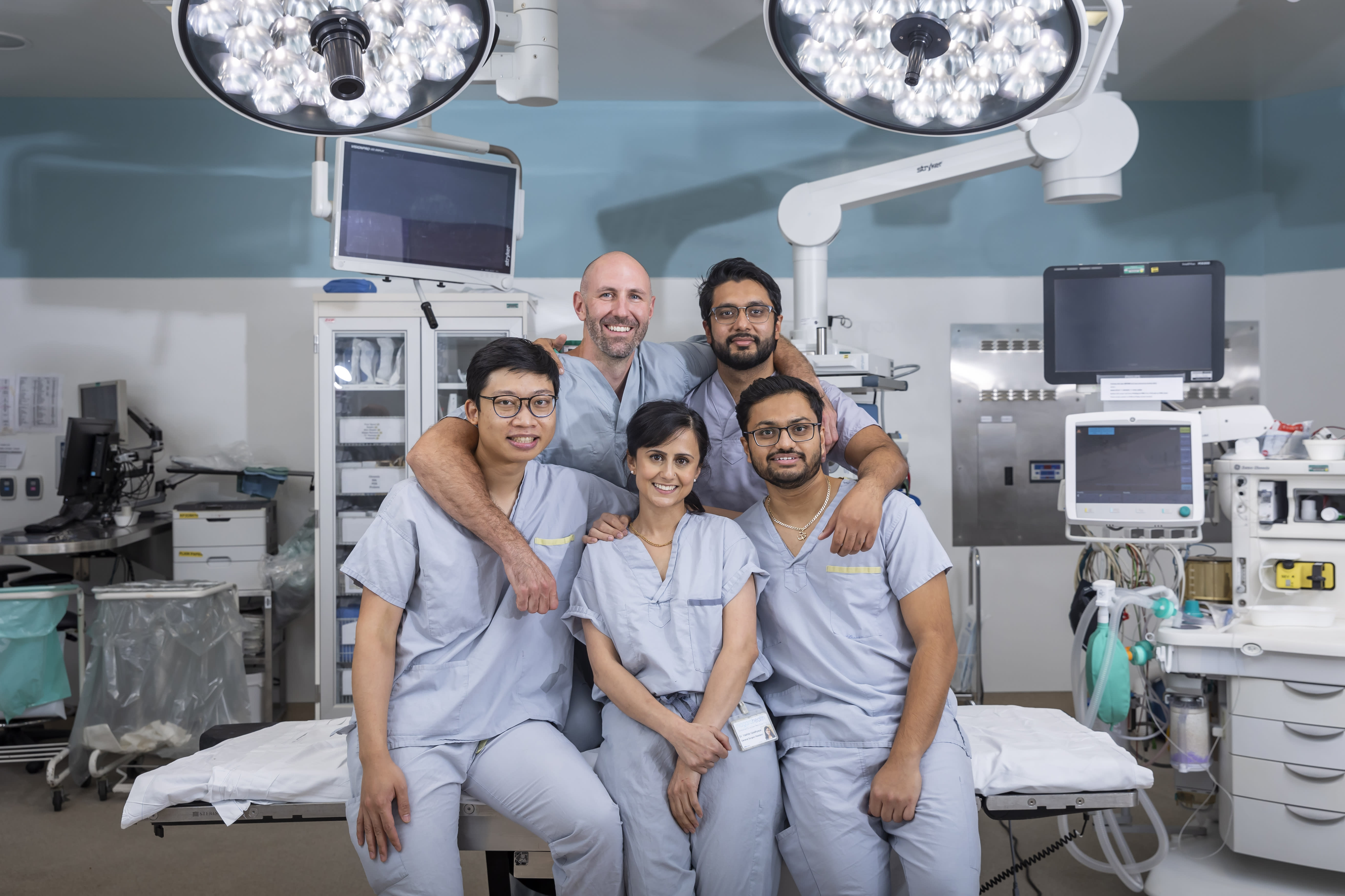 A team of young doctors wearing scrubs gather in a hospital operating room for a photo. They are smiling and surrounded by surgical equipment.