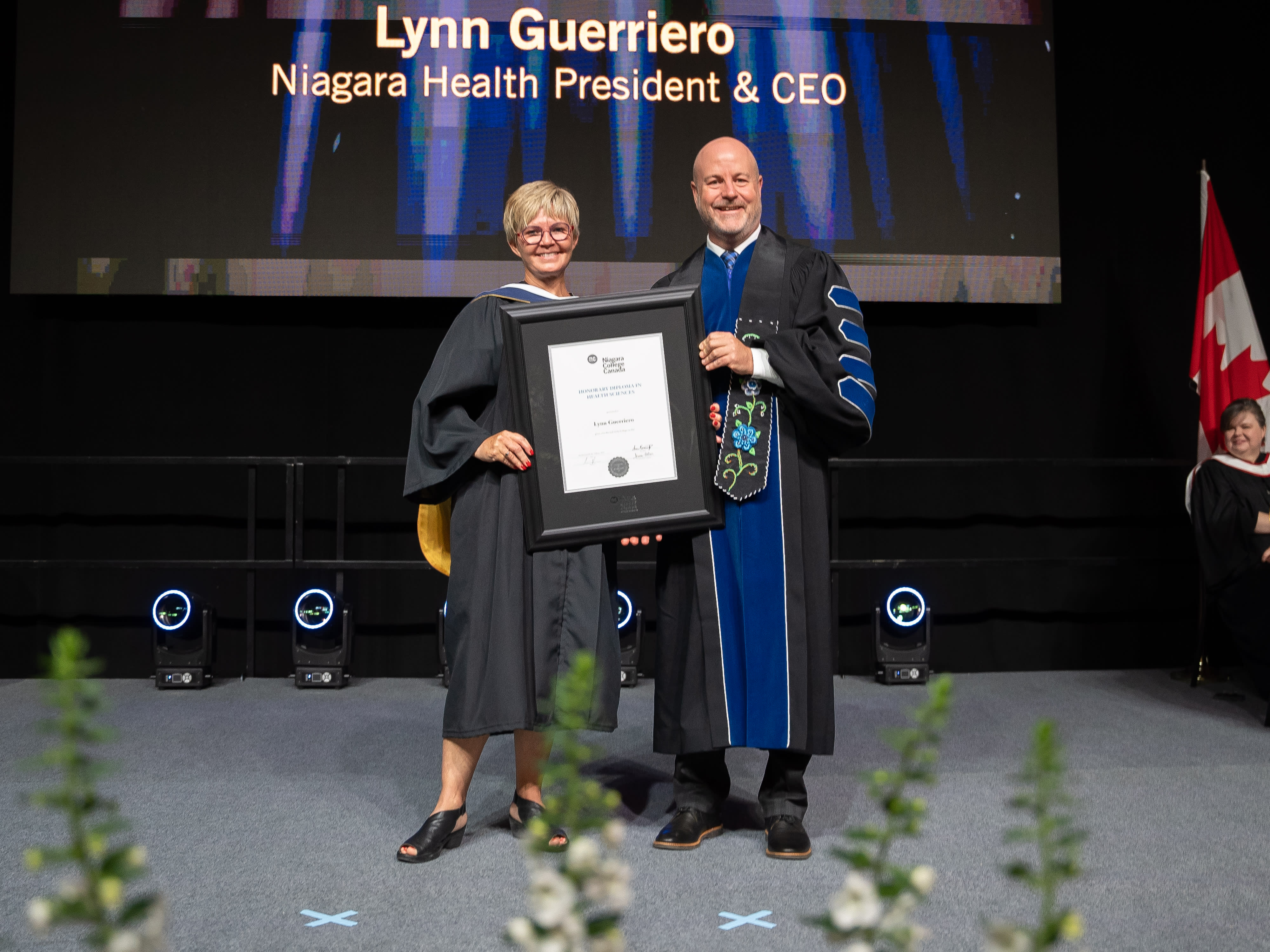 A woman and man stand in graduation gowns on a stage holding a framed diploma between them.