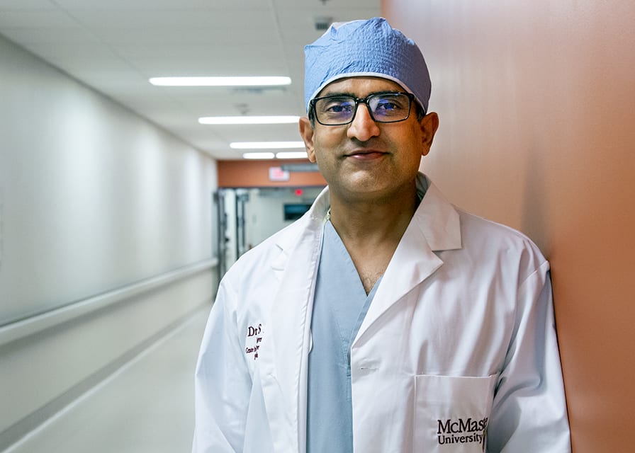 A doctor wearing a white coat, scrubs and cap, leans against a wall in a hospital hallway.