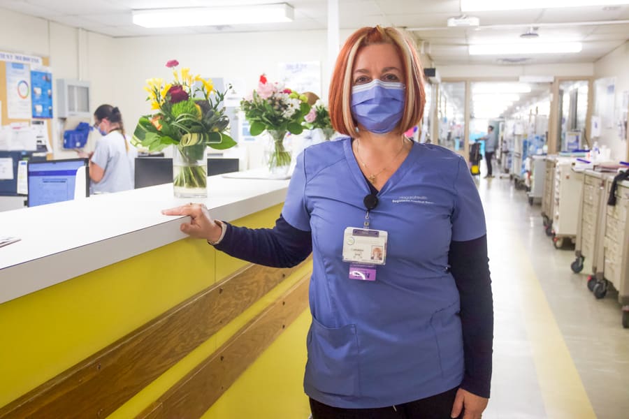 A woman dressed in scrubs and wearing a medical mask stands in the hallway of a busy hospital ward.