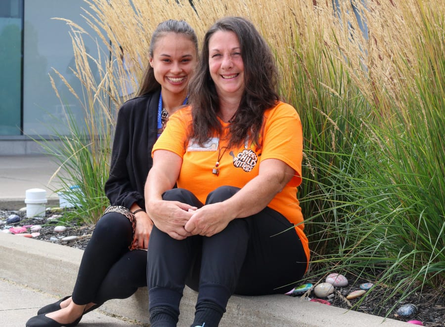 Two Indigneous women sit on the edge of a garden with painted rocks and ornamental grasses. 