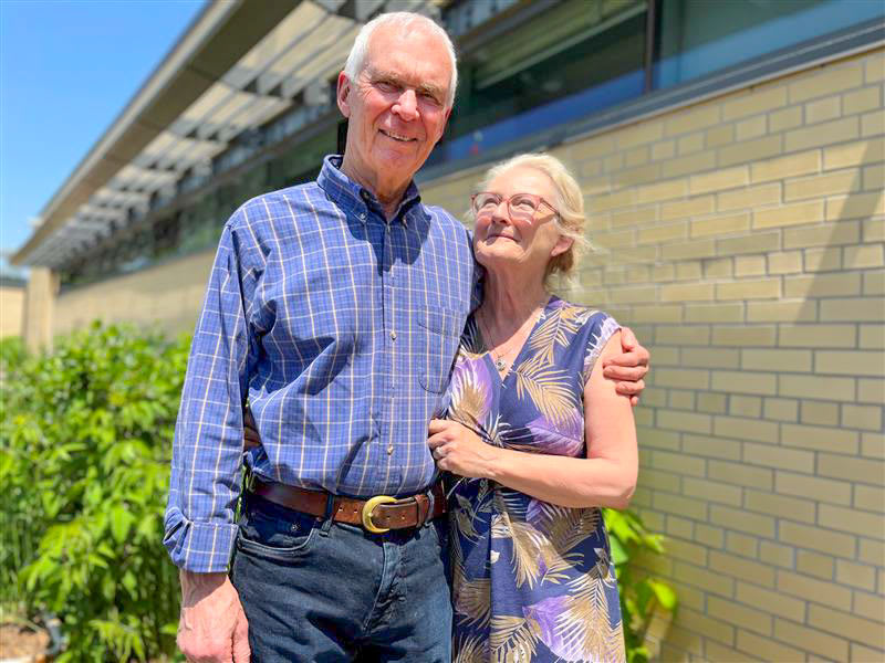 A man and a woman stand with their arms around each other, outside on a sunny day.