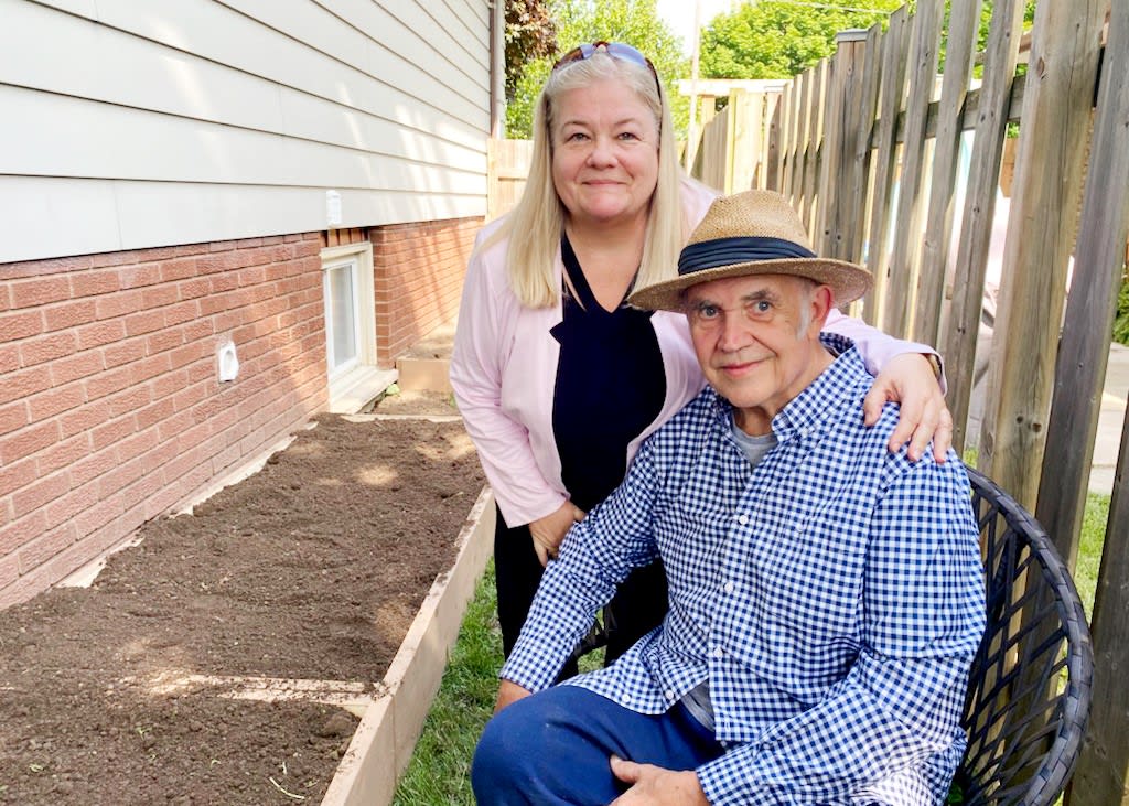 A man sits on his chair in his garden. His wife crouches next to him with her arm around him.