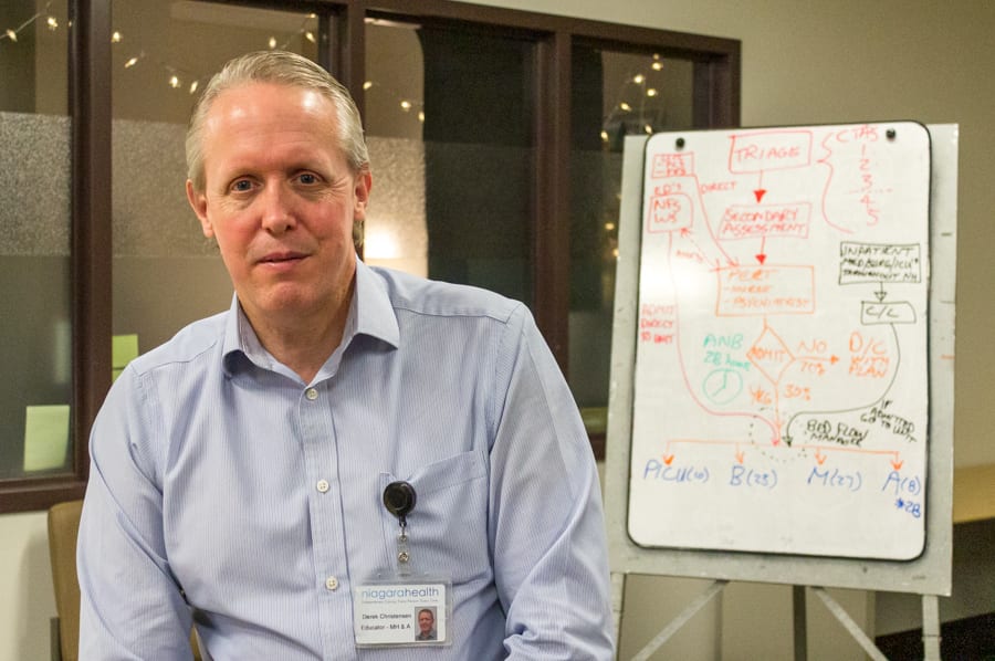 A man in a blue button-up shirt sits with his back toward a white board with a detailed diagram on it.