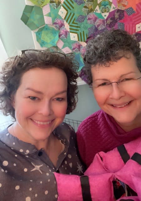 Two women pose with their heads together, holding small cushions in their hands