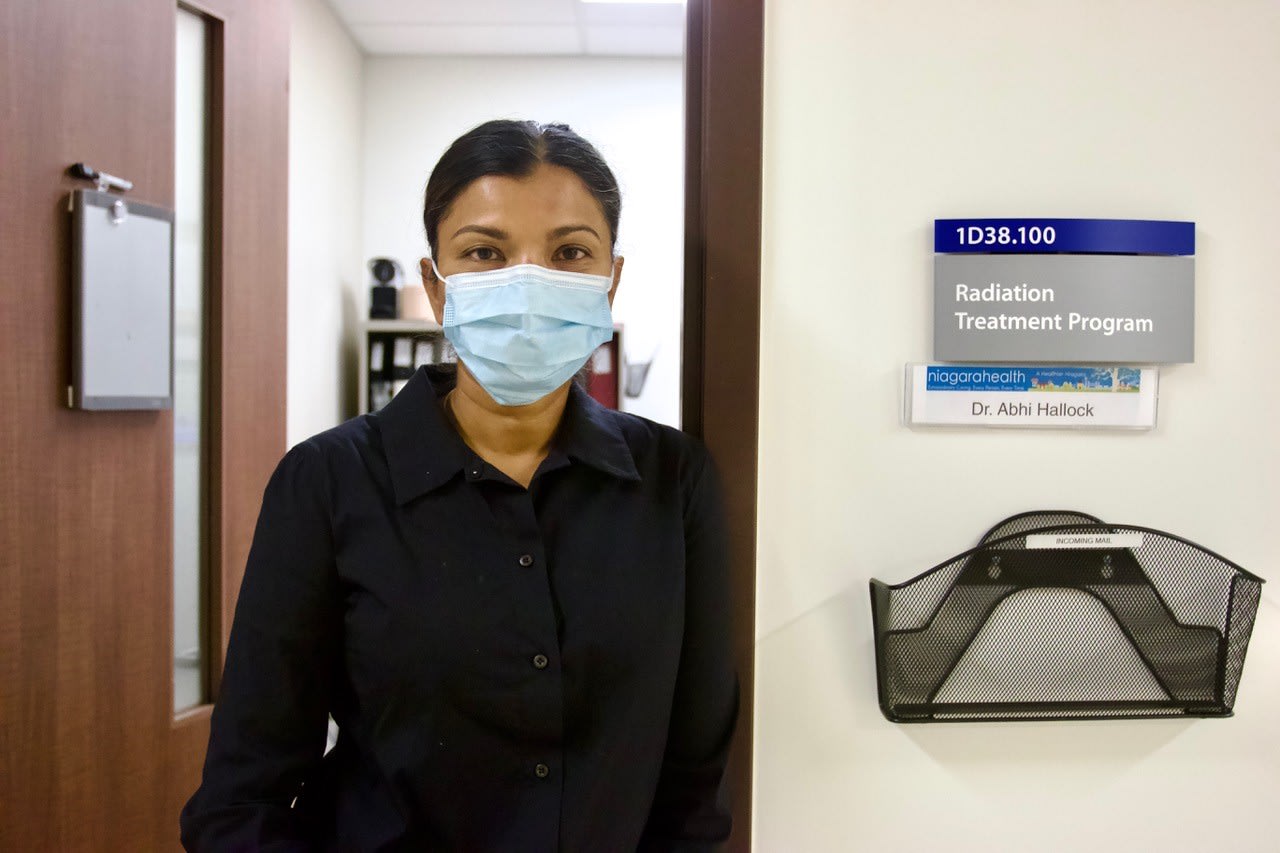 A woman with dark hair and wearing a medical mask stands in the doorway of her office at a hospital.