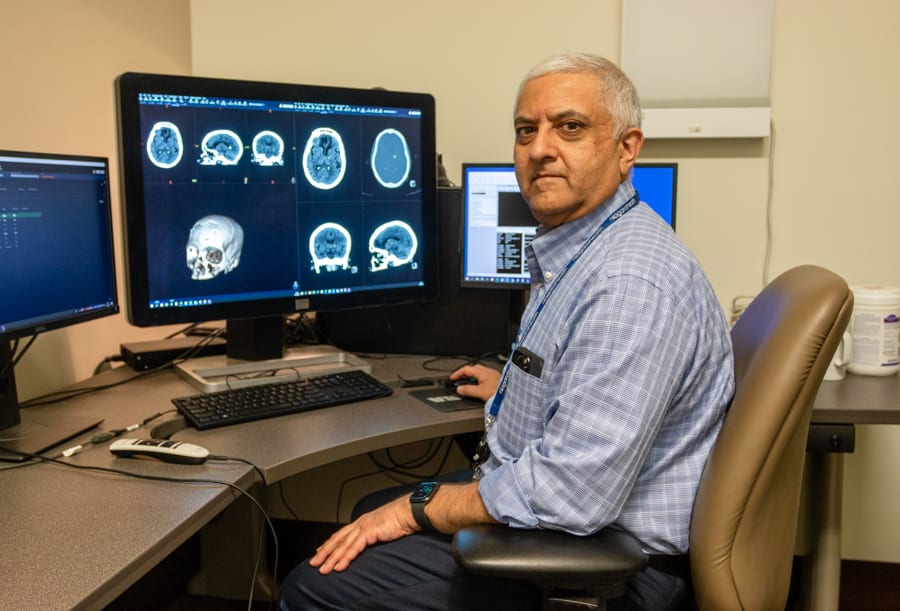 A South Asian man sits at a desk with a computer monitor showing images of a patient's brain scan.