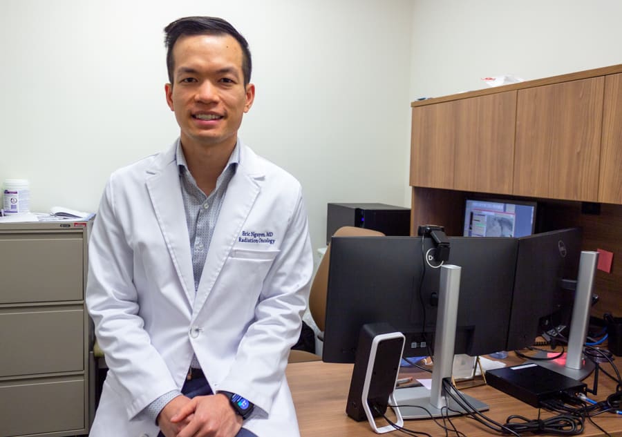 A doctor in a white coat sits on the edge of his desk.