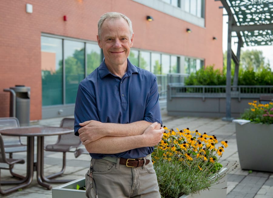 A man with white hair and wearing a blue golf shirt stands in a hospital courtyard smiling for the camera. 