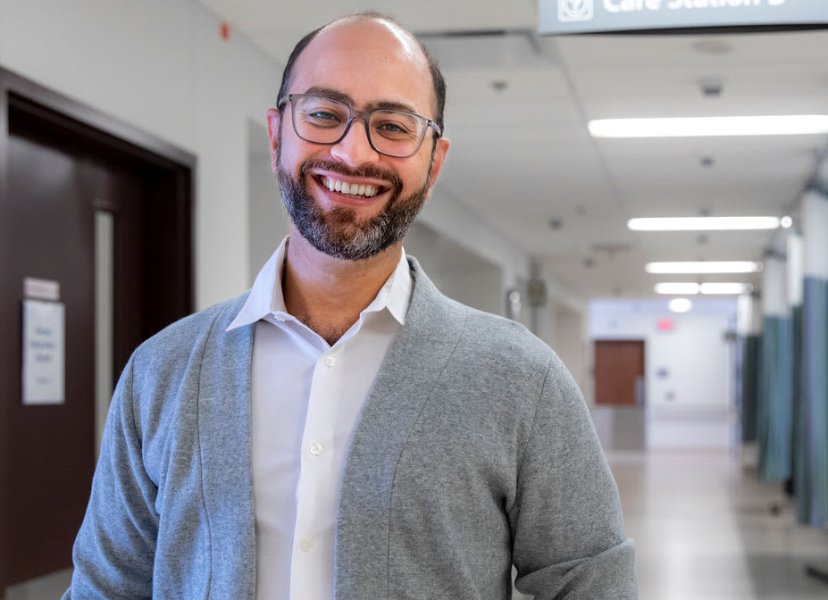 A man wearing a beige cardigan sweater and glasses smiles for the camera