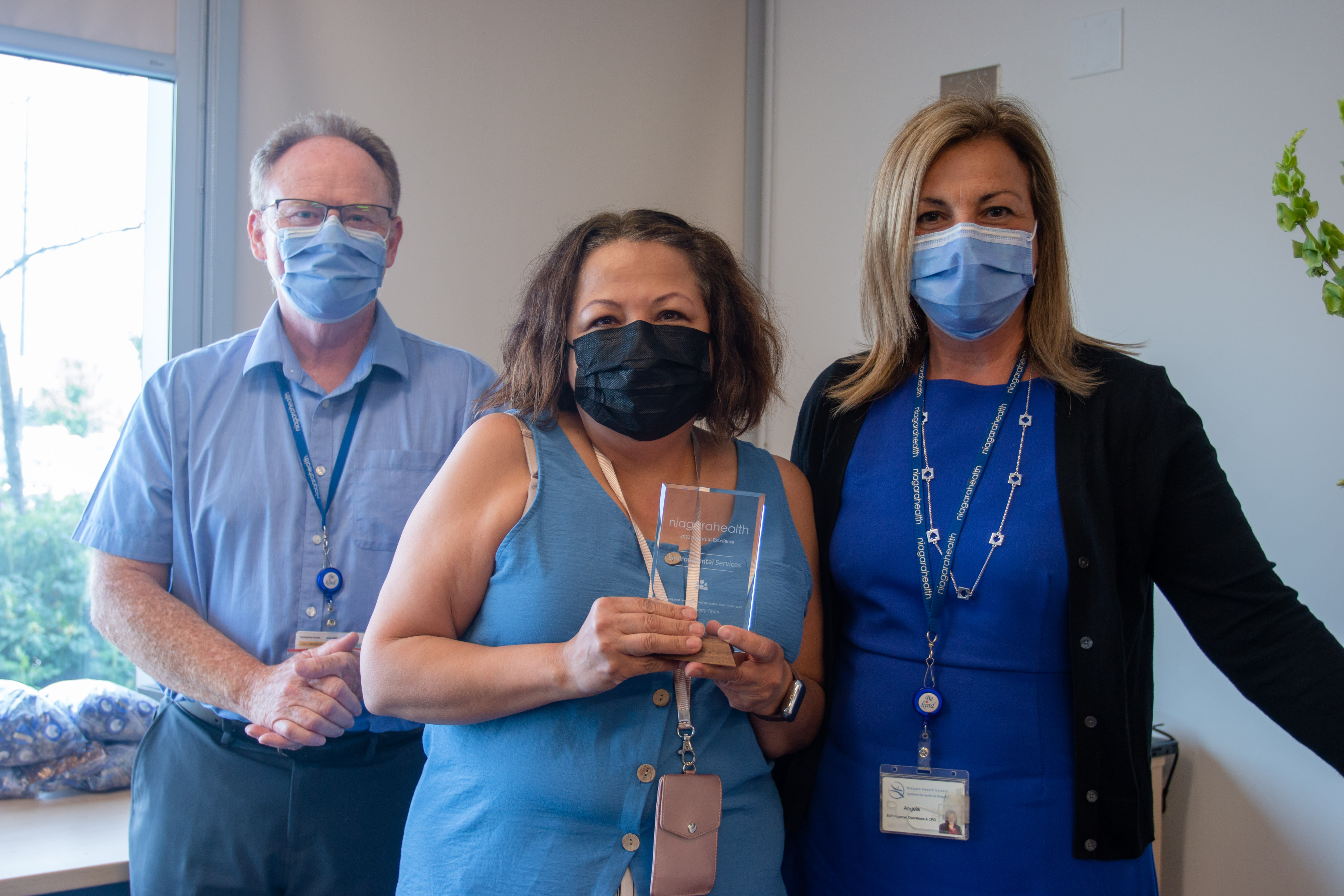Two women and one man stand together. A woman in the middle holds an award. They are all smiling behind their masks.