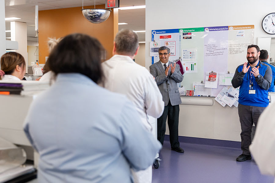 A team of clinicians gather around a huddle board while their leaders clap for them and celebrate their work