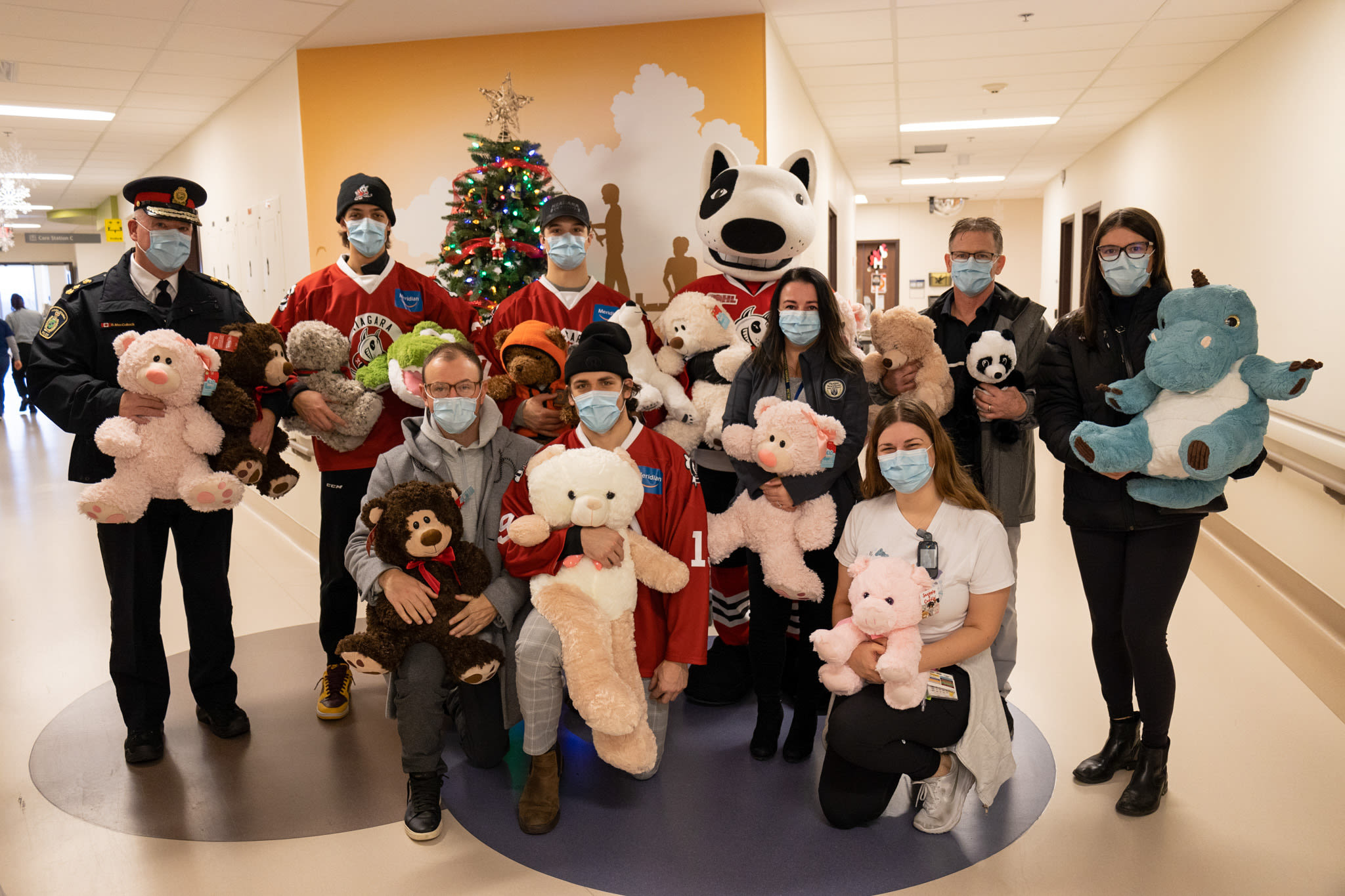 Niagara IceDog players, Chief MacCulloch and representatives from Victim Services Niagara standing in front of a Christmas tree holding teddy bears in the Children's Health Unit at Niagara Health 