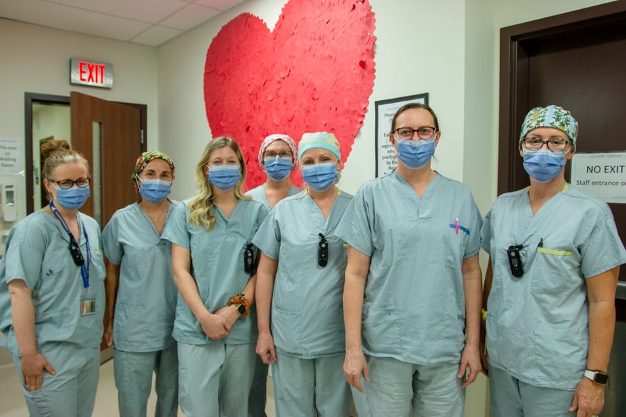 A team of nursing and medical staff wearing scrubs and masks stand against the backdrop of a giant heart made up of smaller cutout hearts