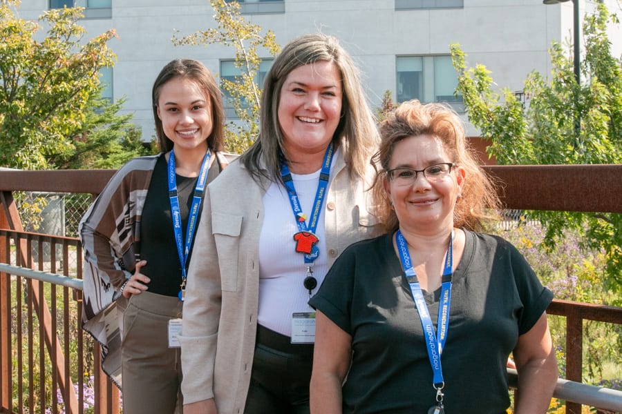 A trio of Indigenous women stand outside a hospital on a sunny day.