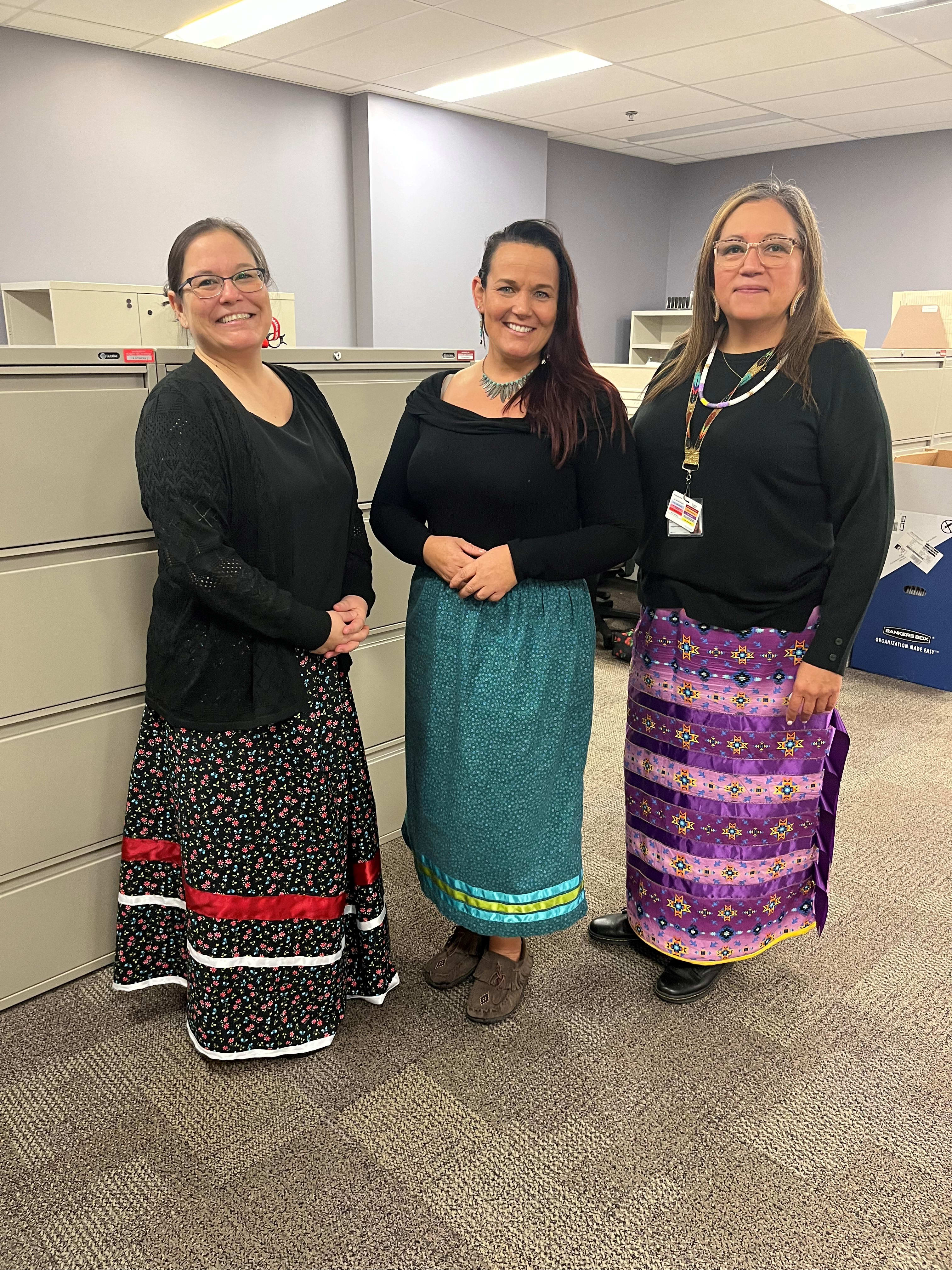 Three Indigenous women gather for a photo in an office. They are wearing ribbon skirts, which are worn for celebrations and significant events. 