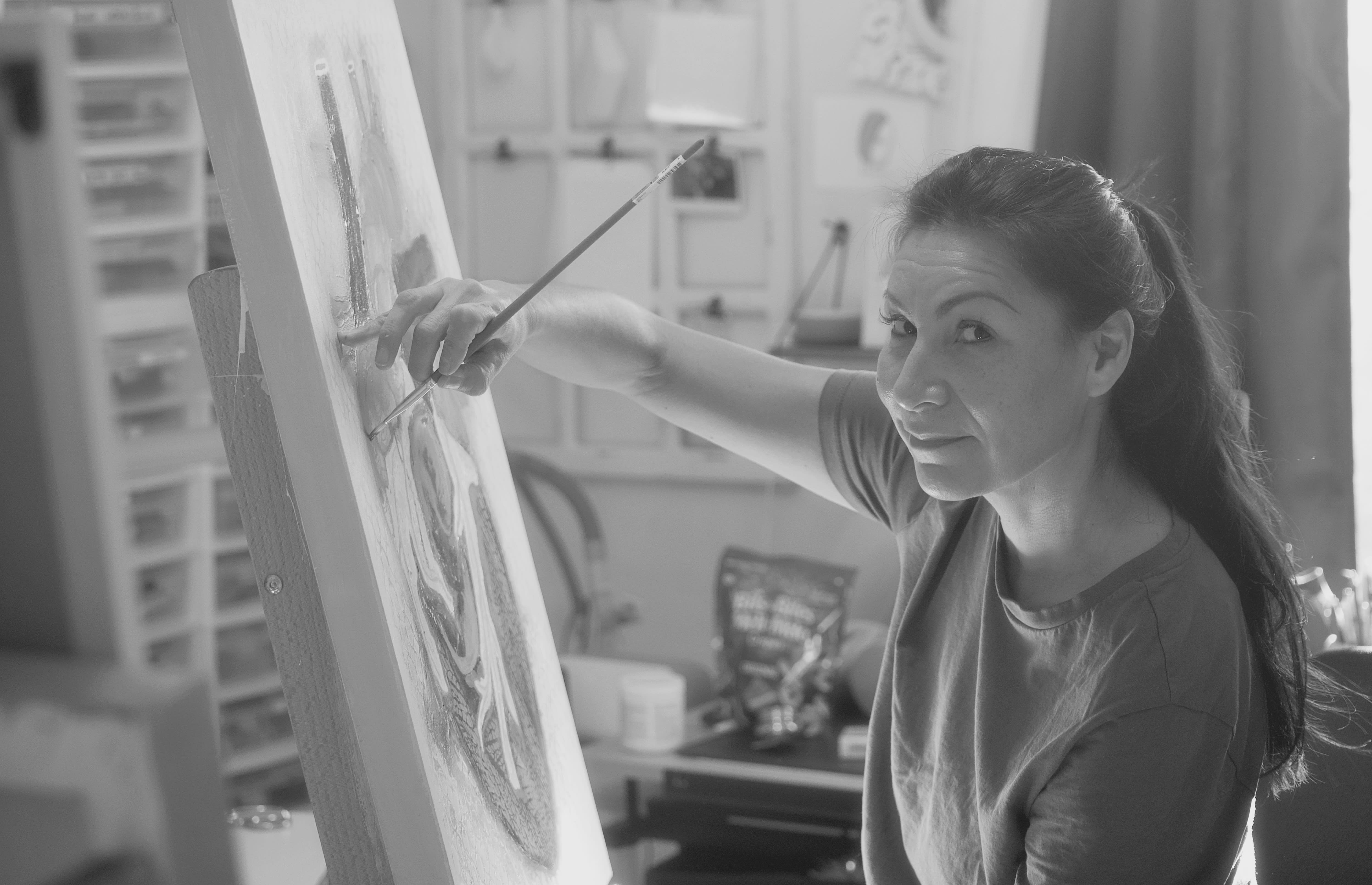 A black and white photo of a woman looking at the camera while she sits at an easel painting.