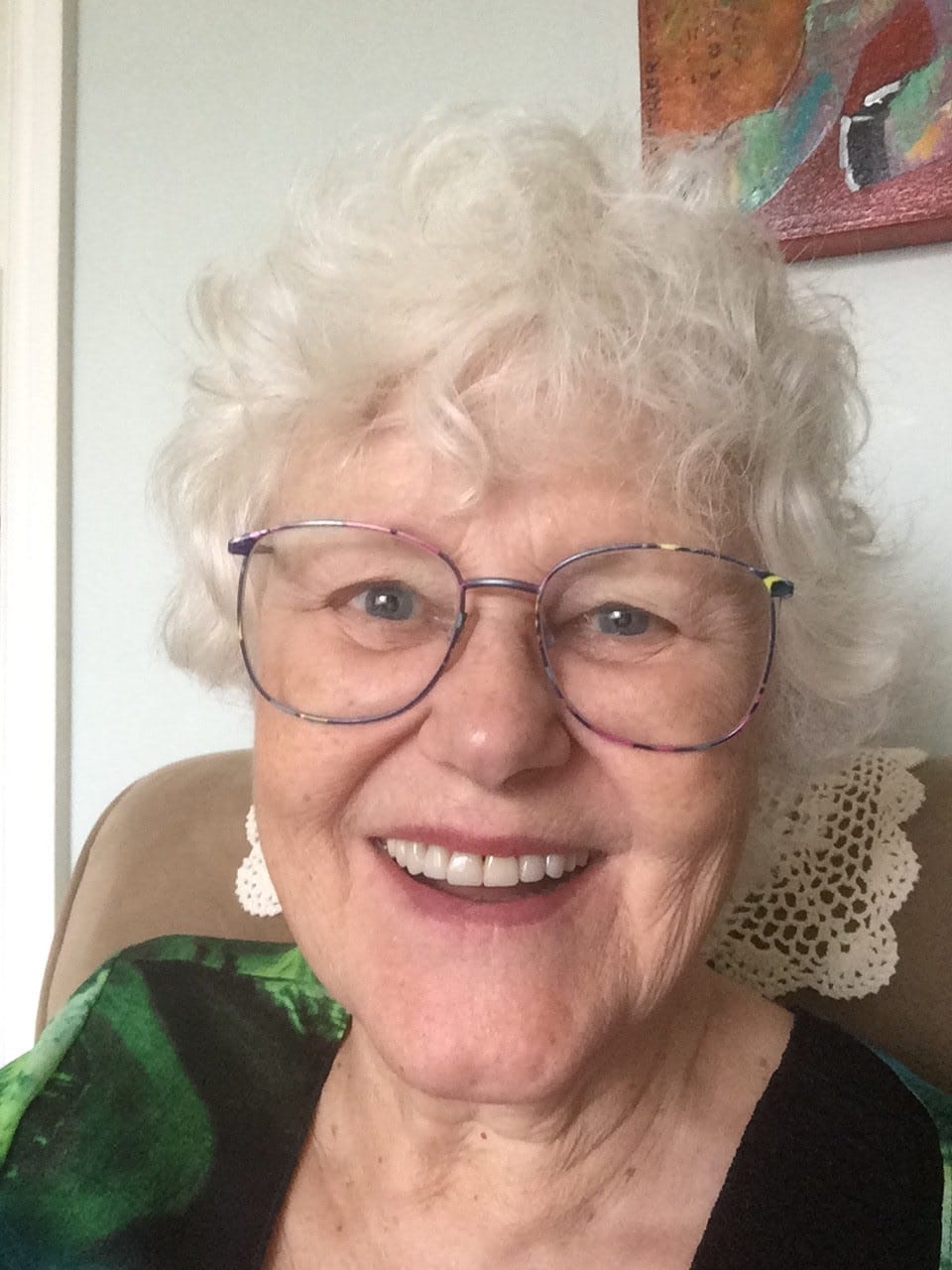 An elderly woman with glasses and curly hair smiles brightly at the camera