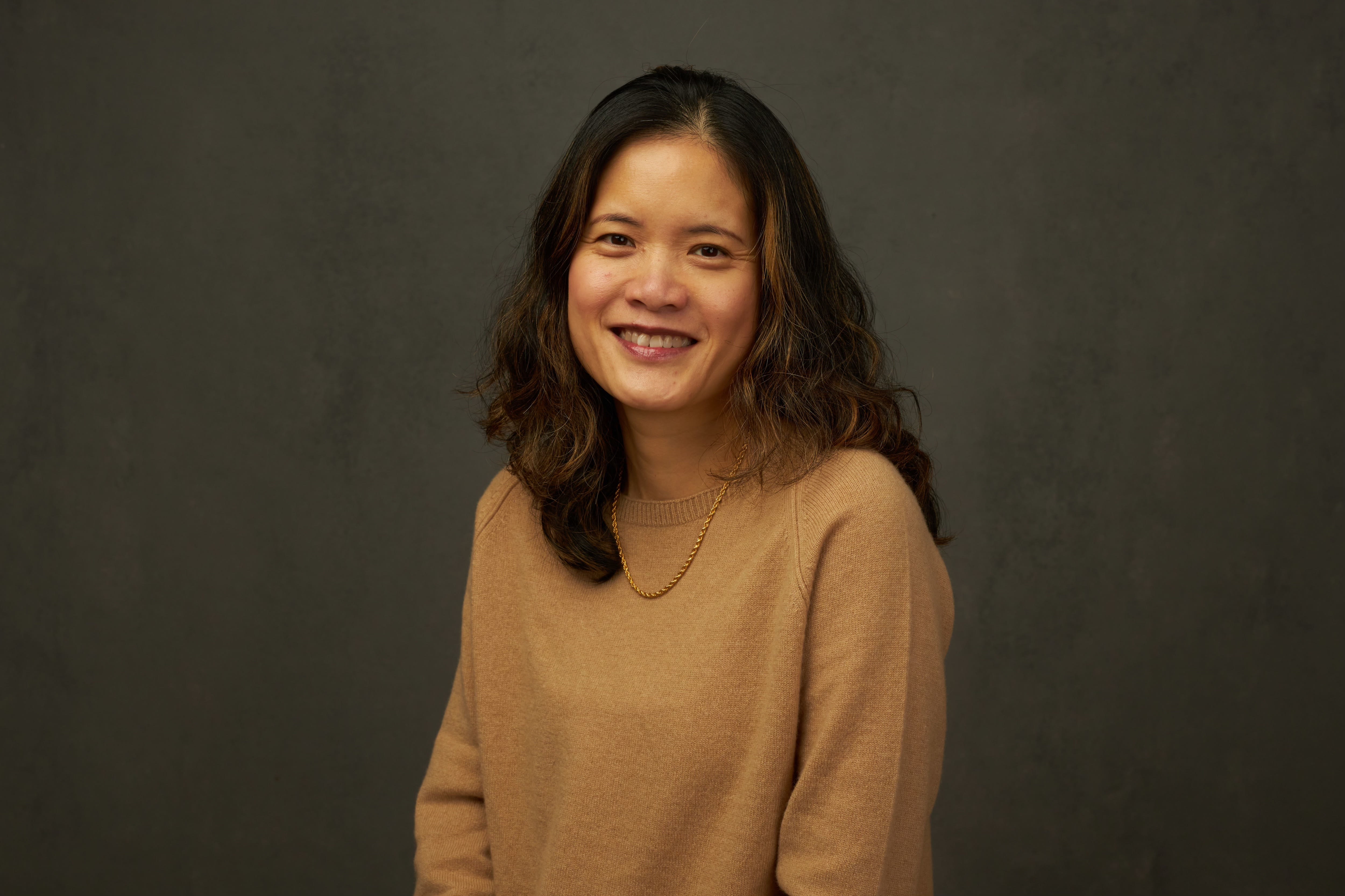An Asian woman wearing a gold-coloured sweater poses for a portrait against a brown background