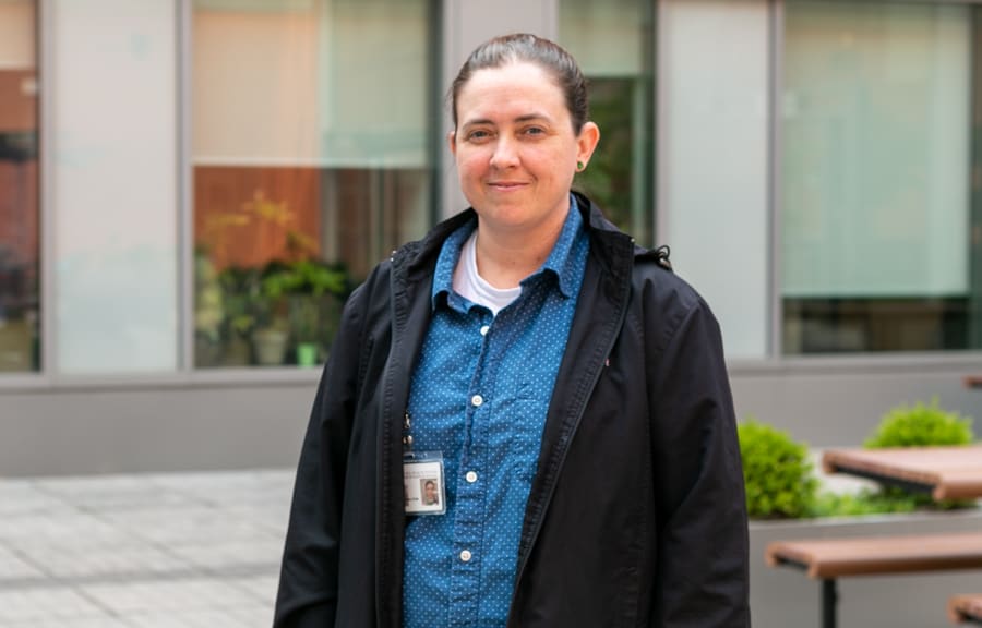 A person stands outside a hospital smiling for the camera. They are wearing a blue shirt and black coat,and wearing their workplace ID.