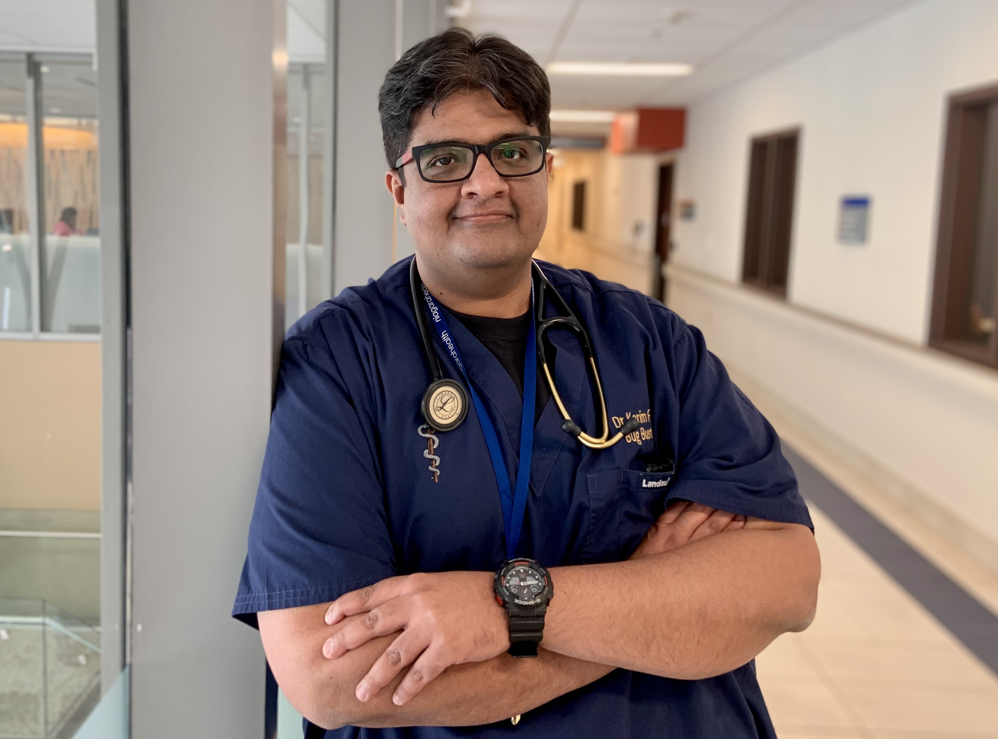 A Brown doctor stands in blue scrubs in a hospital hallway, posing for a photo. His arms are crossed.