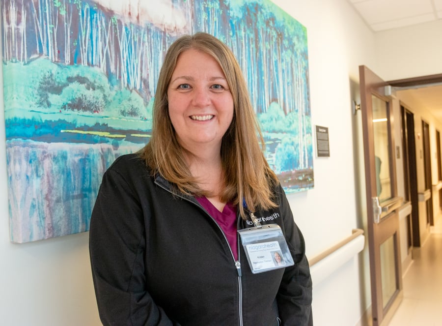 A woman in scrubs and a black sweater stands against an abstract painting in a hospital hallway