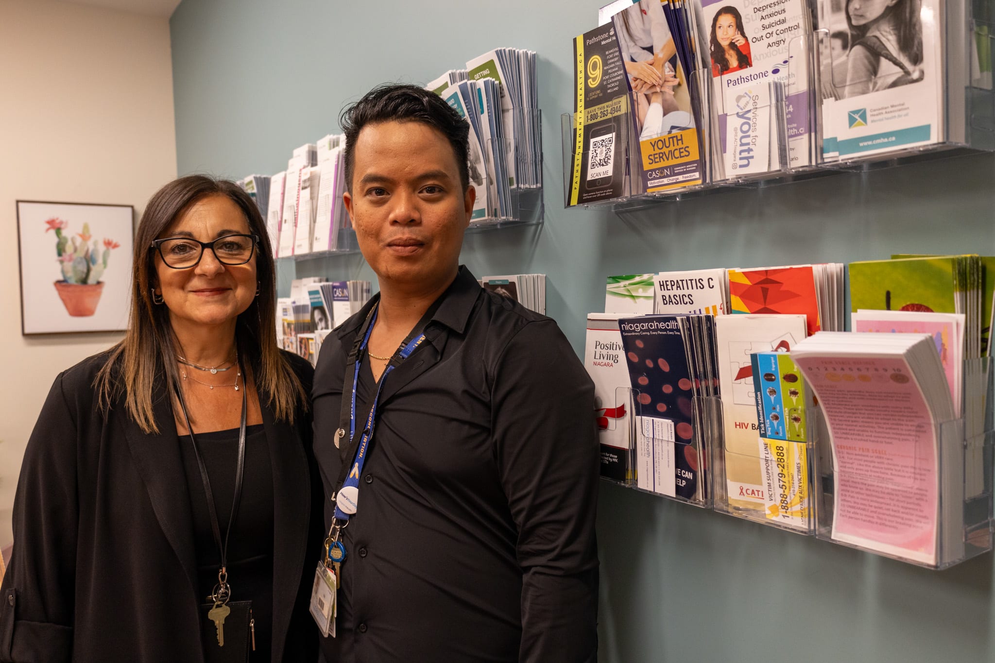 A woman and a man standing side-by-side in front of a wall of community program brochures 