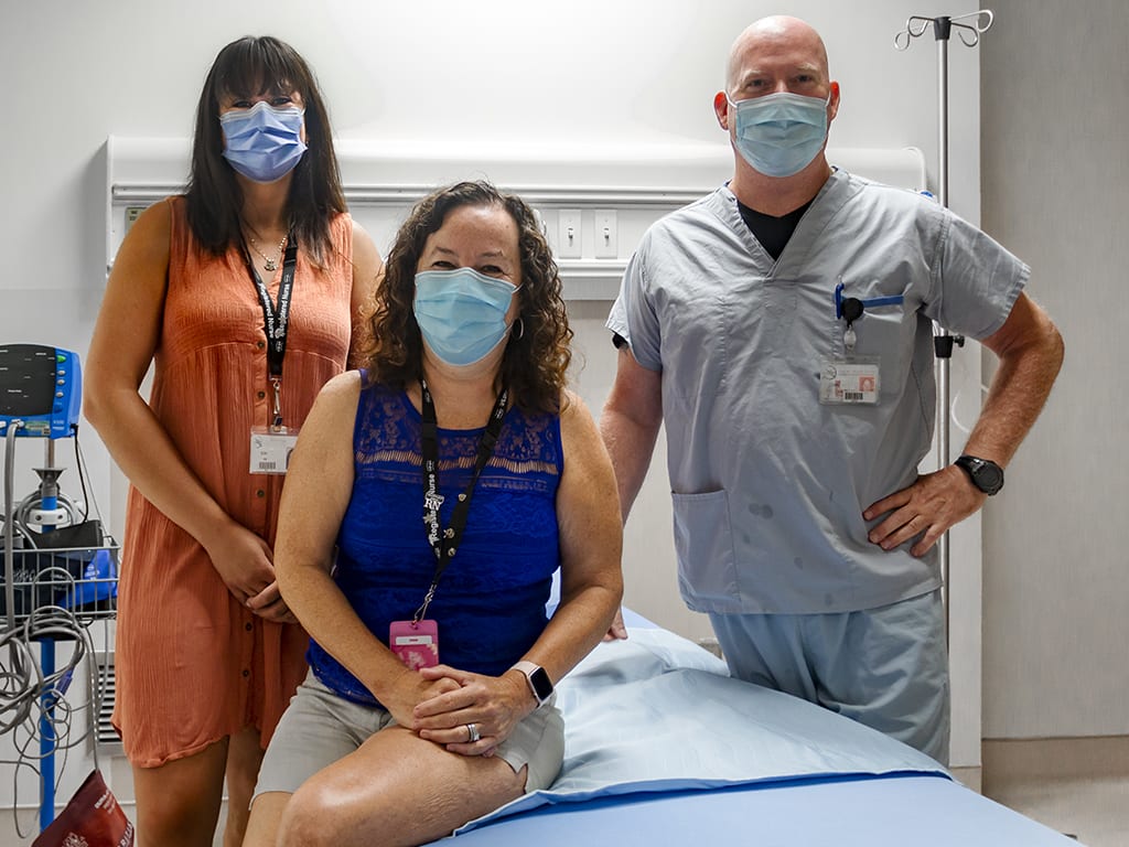 Three people gather around an empty hospital bed inside a patient recovery room. 
