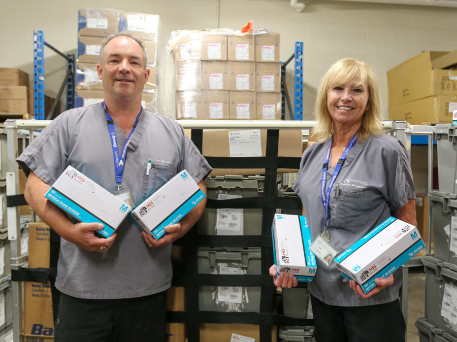 Two people dressed in scrubs stand in a warehouse holding boxes of medical gloves