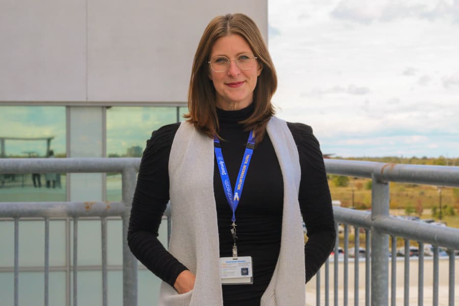 A woman stands on a balcony outside a hospital, smiling for the camera