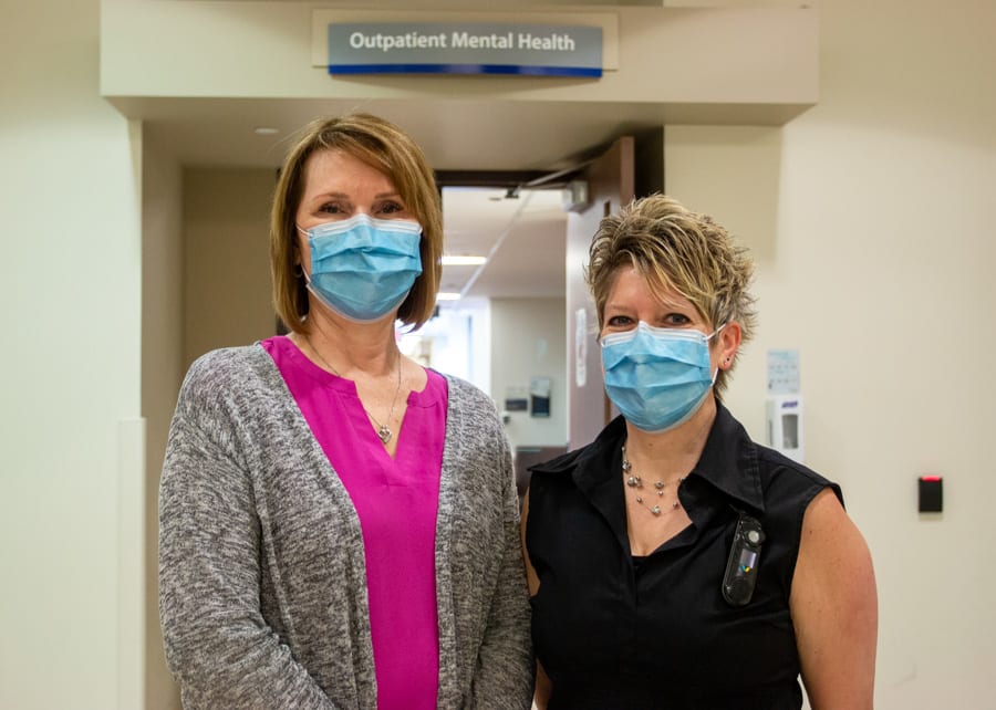 Two women wearing medical masks stand under a sign that reads Outpatient Mental Health in a hospital hallway