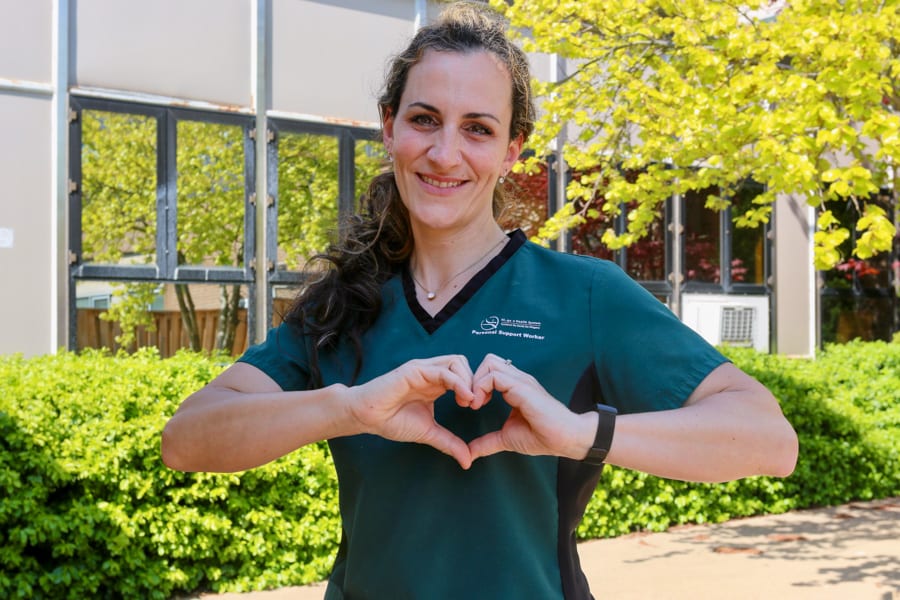 A woman in green scrubs stands outside a hospital and makes a heart with her hands as she smiles for the camera.