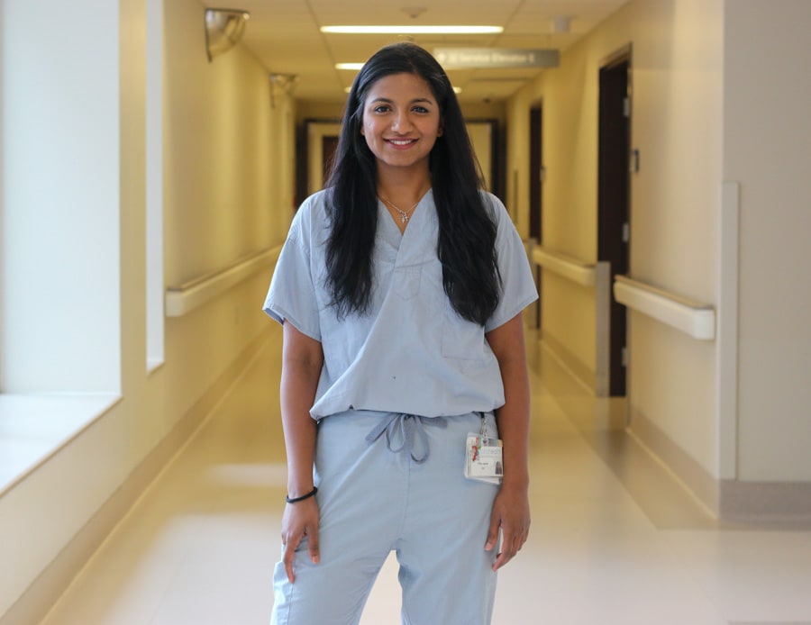 A woman wearing scrubs stands in the hallway of a hospital, smiling for the camera
