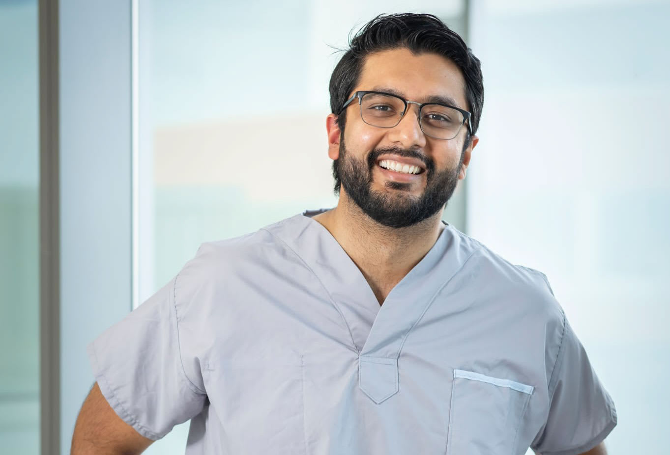 A Brown man dressed in scrubs and wearing glasses smiles for the camera