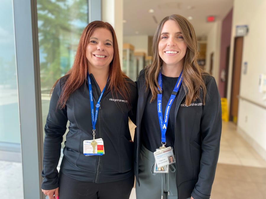 Two women wearing black Niagara Health zip-up jackets stand arm in arm smiling for the camera in a hospital hallway