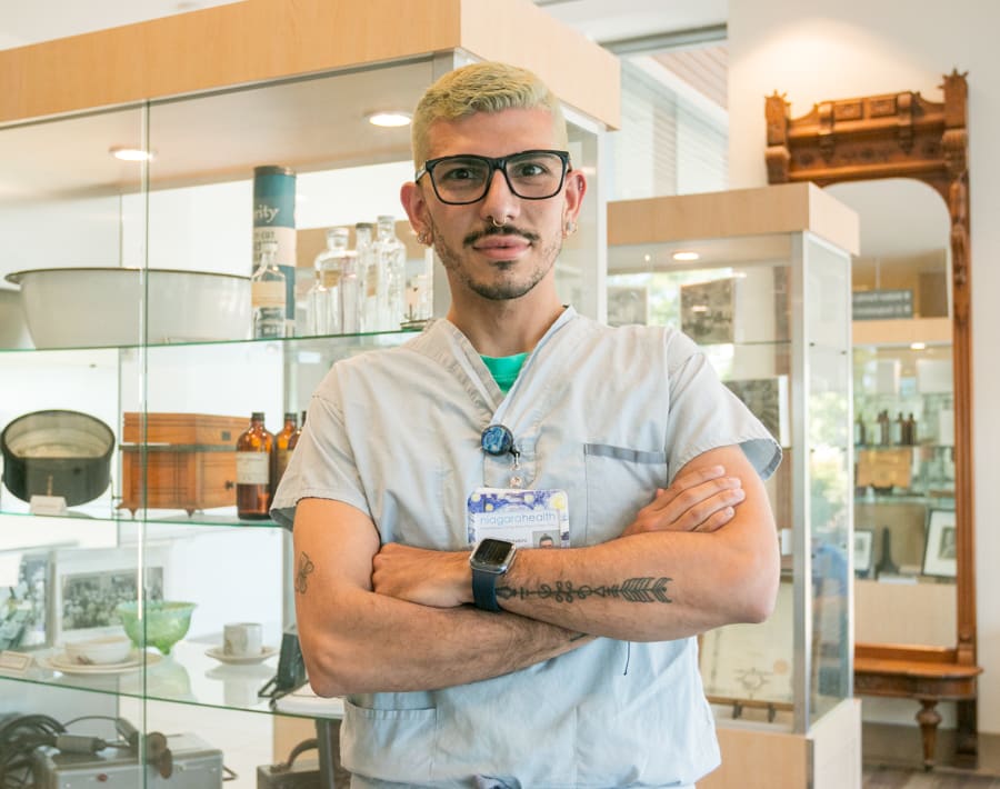 A Latino man with blond hair, 5:00 shadow and glasses wears scrubs and stands with his