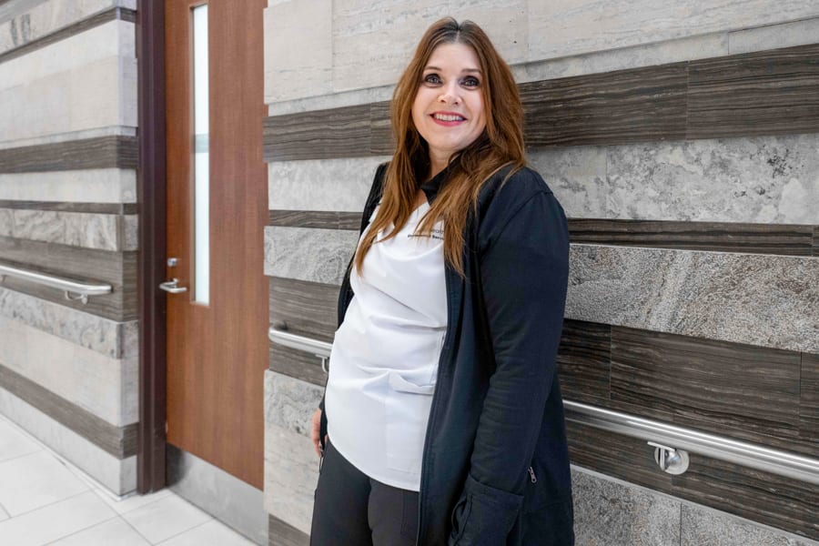 A woman wearing a black sweater and white scrubs stand with her back against a granite wall