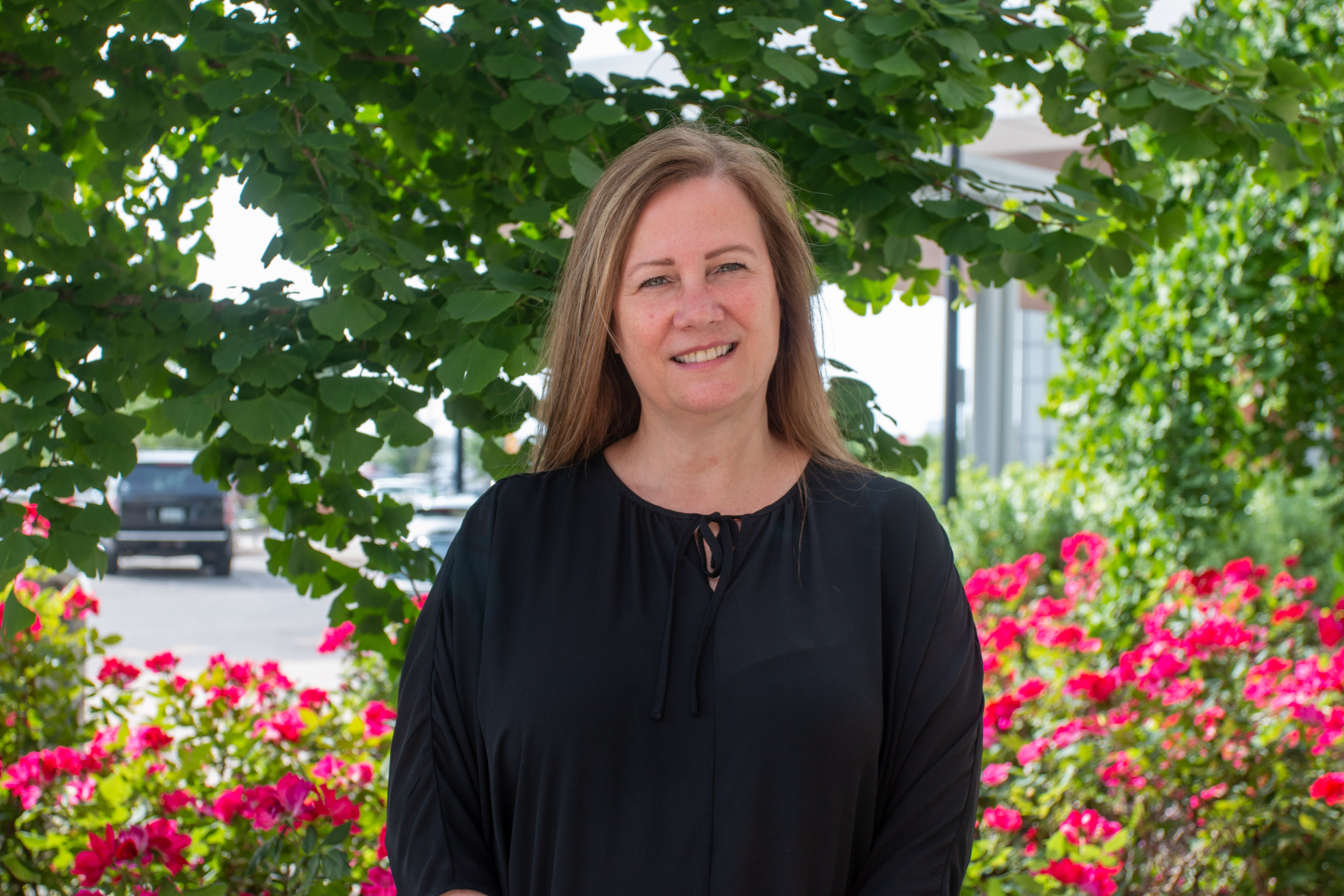 A woman dressed in black stands against a rose garden and smiles slightly for the camera.