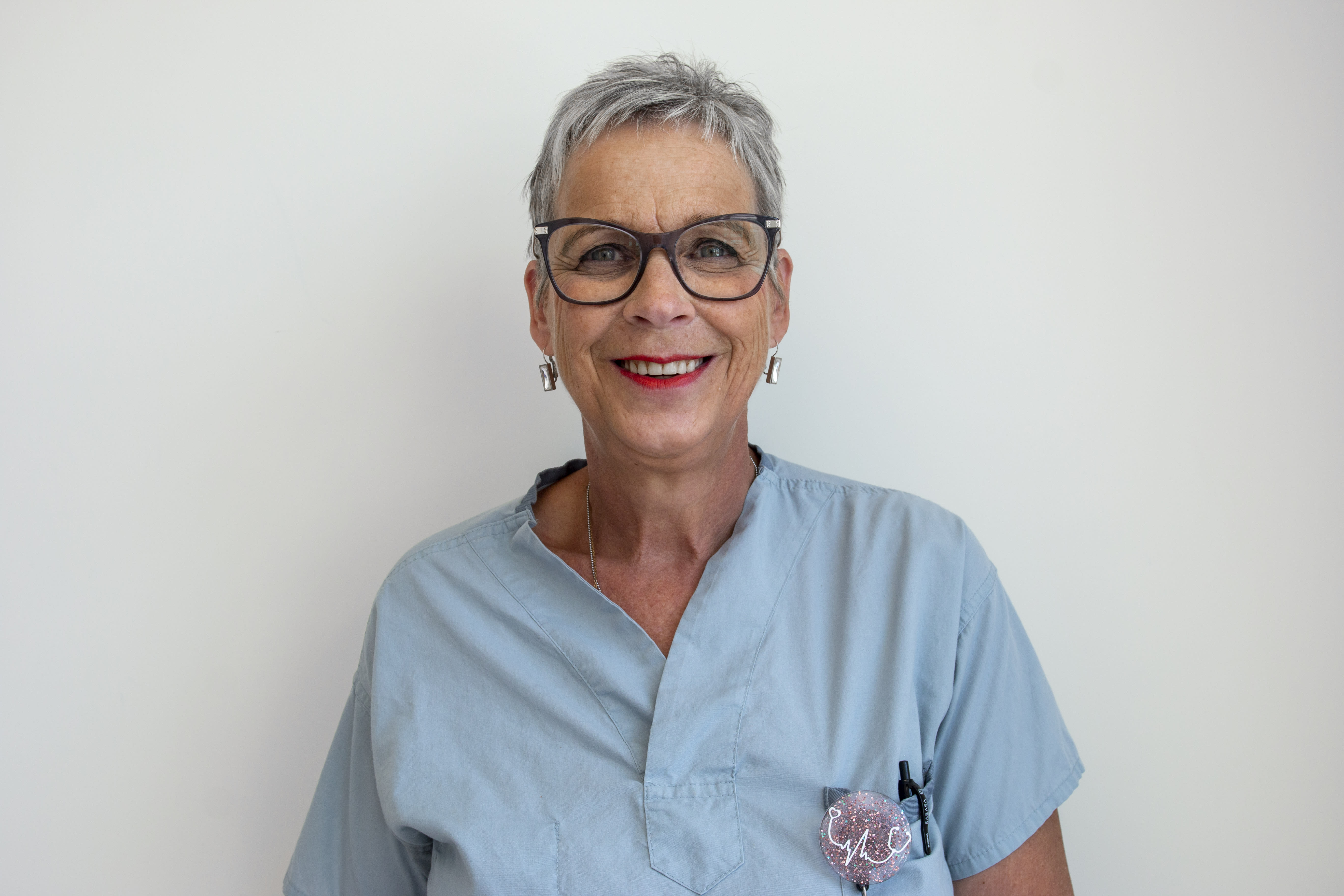 A woman smiles brightly for the camera. She is wearing scrubs and stands against a plain background.