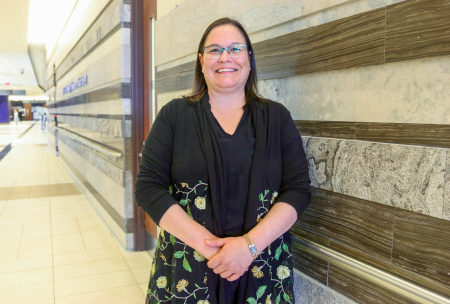 An Indigenous woman with shoulder length hair and glasses leans against a granite wall and smiles for the camera.