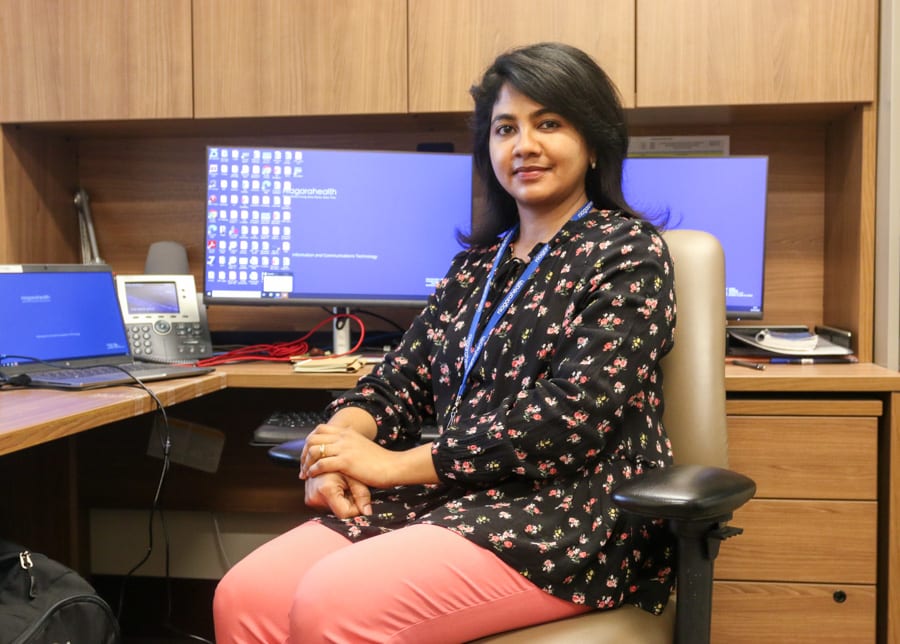 A South Asian woman sits in a chair near her desk in a hospital office. She is looking at the camera.