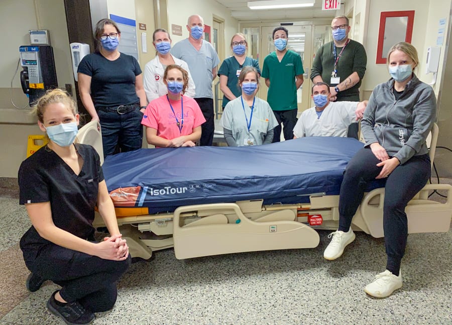 A group of people dressed in scrubs gather around a hospital bed in the hallway of a hospital